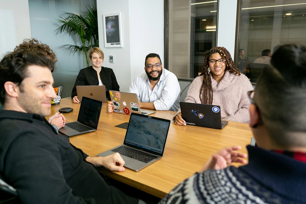 team meeting with laptops 1000×667 Six people sit around a conference table with laptops, smiling and engaged in a meeting in a modern office setting.