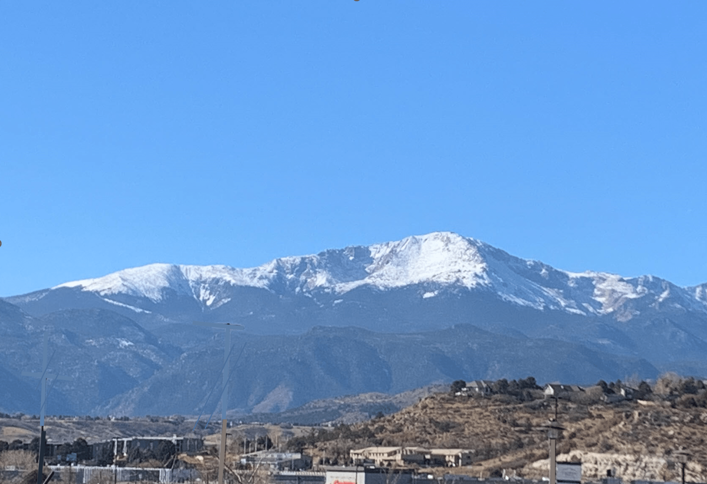 Pikes Peak – touched up A scenic landscape featuring snow-capped mountains under a clear blue sky. There are scattered buildings and a few streetlights in the foreground, with green vegetation covering the hills at the base of the mountains.